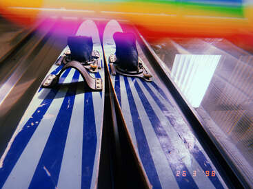 Esta imagen muestra un primer plano de dos esquís a rayas azules y blancas, llevados en las fijaciones, con una fuga de luz de colores y un sello de fecha retro.