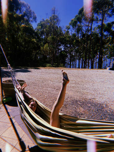 Una persona se relaja juguetonamente en una hamaca a rayas, con una pierna levantada y los brazos extendidos, rodeada de árboles y cielo azul en un día soleado al aire libre.
