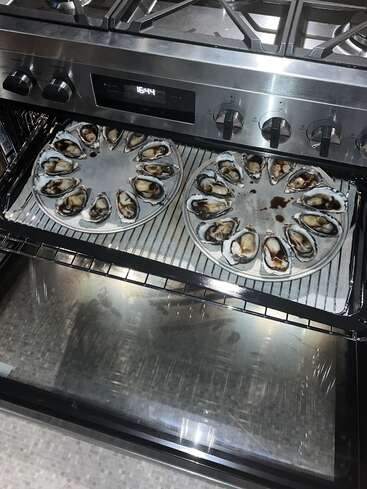 Dos bandejas llenas de ostras frescas, cada una cubierta con una salsa oscura, están dispuestas dentro de un moderno horno, listas para ser cocinadas u horneadas.