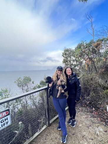 Dos mujeres de pie junto a la valla de un acantilado, cada una con un perro pequeño en la mano. El cielo está parcialmente nublado, con el océano y las islas distantes visibles en el fondo.