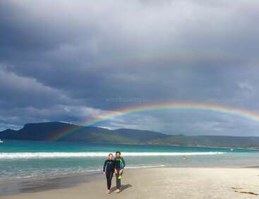 Dos personas en traje de neopreno caminan por una playa de arena bajo un hermoso arco iris doble, con las olas turquesas del océano y un dramático cielo nublado de fondo.