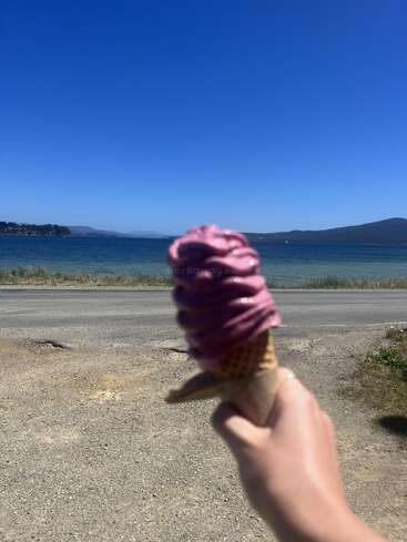 Una mano sostiene un cucurucho de helado morado, con un cielo azul despejado, un lago tranquilo y colinas lejanas de fondo en un día soleado.