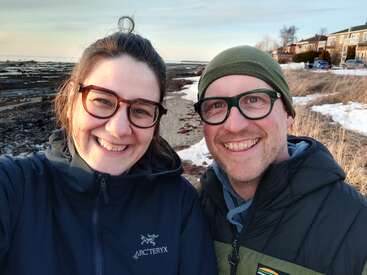 Um casal sorridente, de óculos e bem agasalhado, está ao ar livre perto de uma costa rochosa e coberta de neve ao pôr do sol. No fundo, dá pra ver casas e grama seca.