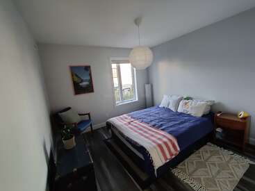 Este quarto aconchegante tem uma cama de casal com roupa de cama azul, uma manta listrada, uma mesinha de cabeceira, uma lanterna de papel, uma cadeira, um baú, uma janela, um tapete e uma decoração de parede.