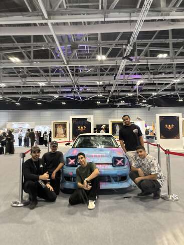 Fünf Männer posieren bei einer Indoor-Ausstellung vor einem individuell gestalteten blauen Auto mit rosa Graffiti, im Hintergrund sind gerahmte Kunstwerke und Menschen zu sehen.