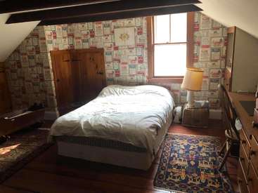 The image depicts a cozy bedroom with a bed, wooden furniture, and a window, featuring a vibrant rug and wallpaper with a unique design.