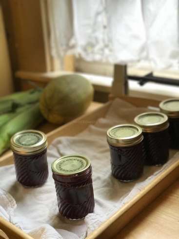 The image depicts four jars of dark purple jam on a wooden tray, with a cantaloupe and green vegetables in the background, set against a window with white curtains.