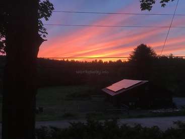 The image depicts a serene sunset scene with a house featuring solar panels, surrounded by trees and a road, set against a vibrant sky with pink and orange hues.