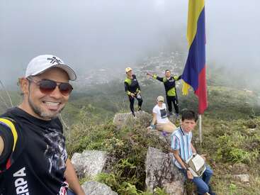 Cinco pessoas curtem o topo de uma montanha enevoada ao lado de uma grande bandeira colombiana. Sorridentes, elas posam para uma selfie, cercadas por uma vegetação exuberante e vistas distantes da cidade.