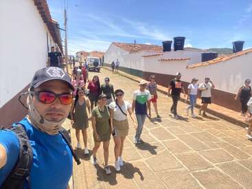 Um grupo de pessoas caminha por uma rua ensolarada de paralelepípedos, ladeada por casas brancas e telhados vermelhos. Elas parecem animadas, aproveitando um dia bonito juntas.