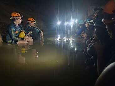 Um grupo de pessoas usando capacetes e lanternas de cabeça está sentado na água rasa da caverna, cercado pela escuridão; as luzes refletem na superfície da água, criando uma atmosfera de aventura.