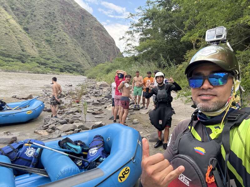 Eine Gruppe von Leuten bereitet sich auf eine Rafting-Tour auf einem von Bergen umgebenen Fluss vor. In der Nähe liegen zwei blaue Schlauchboote. Ein Mann macht ein Selfie, lächelt und gestikuliert dabei.