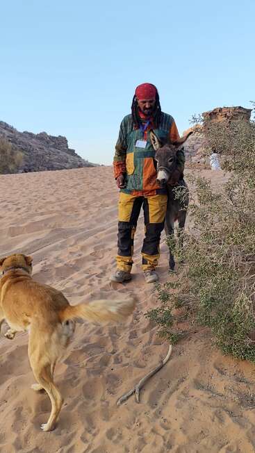 Un homme portant des vêtements colorés et un bandana rouge se tient dans le désert avec un âne, tandis qu'un chien brun court près des buissons et des collines rocheuses.