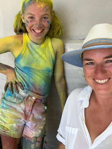 Un niño sonriente, cubierto de pintura de colores, está junto a una mujer con una camisa blanca y un sombrero para el sol. Ambos están felices, disfrutando de una actividad creativa y un poco desordenada.