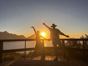 Dos personas posan alegres en una terraza de madera al atardecer, recortadas contra una impresionante vista del océano y las montañas, irradiando felicidad y libertad en medio de la belleza de la naturaleza.