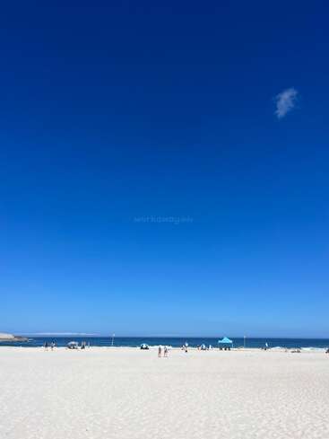Una playa de arena inmaculada bajo un cielo azul inmenso y despejado, con algunos bañistas, sombrillas y una única tienda azul cerca de las suaves olas del mar.