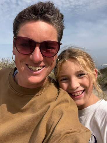 A woman wearing sunglasses and a brown sweater smiles with a young blonde girl in a white shirt. They are outdoors on a bright, sunny day.