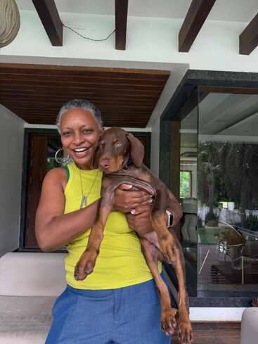 Eine fröhliche Frau in einem gelben Tanktop und einer blauen Hose steht draußen und hält mit einem strahlenden Lächeln einen jungen braunen Dobermannwelpen im Arm.