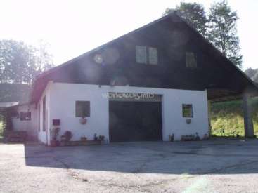 The image depicts a white barn with a black roof and a large black door, featuring a driveway and a grassy area with trees in the background.