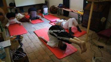 The image depicts a group of individuals practicing yoga on red mats in a room with a brick floor, surrounded by various objects and cords.