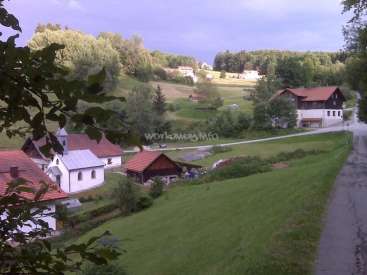 The image depicts a serene rural landscape, featuring a church, houses, and a road, set against a backdrop of lush greenery and a cloudy sky.