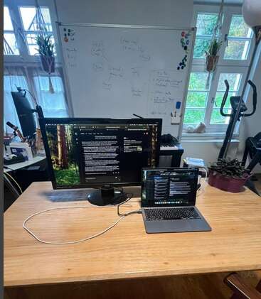 A workspace with a wooden desk, laptop, large monitor, whiteboard with notes, windows, plants, mug, exercise bike, and various office supplies in the background.
