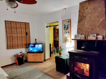 A cozy living room features a lit wood stove, small TV, bamboo blinds, framed artworks, lamp, plants, stone fireplace, and a doorway to another room.
