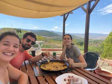 Three friends enjoy a sunny outdoor meal under a canopy, sharing a large pan of paella with mountains and green countryside in the beautiful background. Smiling faces.