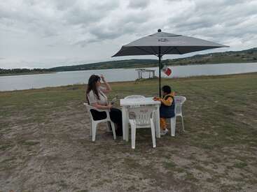 Uma mulher e uma criança estão sentadas a uma mesa branca com um grande guarda-sol à beira de um lago, rodeadas por grama, sob um céu nublado. Há uma bandeira mexicana pendurada.