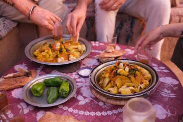 Trois personnes partagent un repas composé de légumes colorés servis dans des plats traditionnels, qu'elles mangent avec les mains. Du pain frais, des poivrons verts et des verres à thé sont disposés sur la table fleurie.