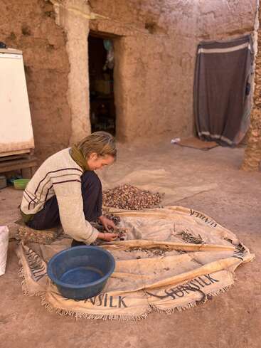 Une personne est assise par terre à l'intérieur et trie des noix ou des graines sur un tissu. On aperçoit en arrière-plan une bassine en plastique, des murs en pierre et un environnement terreux.