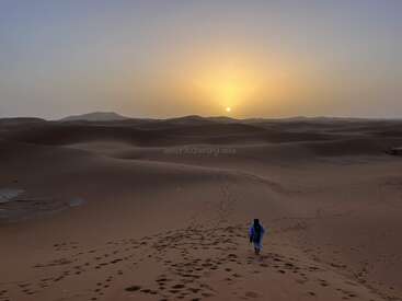 Au coucher du soleil, une silhouette solitaire vêtue de bleu traverse de vastes dunes de sable ondulantes. Des empreintes de pas s'étendent derrière elle, tandis que le soleil doré illumine un paysage désertique paisible.