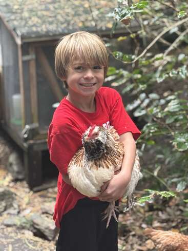 A smiling young boy wearing a red shirt is standing outdoors, happily holding a large chicken. Green foliage and a wooden chicken coop are in the background.
