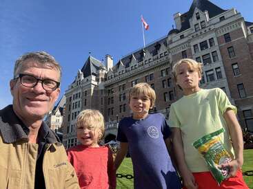 A man and three boys smile in front of a historic, castle-like building with a Canadian flag. The sun is shining, and one boy holds sunflower seeds.