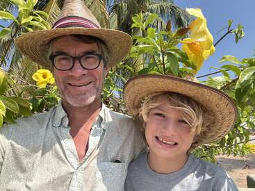 A man and a boy, both wearing straw hats, smile together outdoors. Lush green foliage, yellow flowers, and palm trees create a bright, sunny background.
