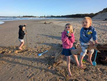 Drei Kinder spielen an einem Sandstrand. Eines gräbt mit einem Stock, ein anderes sitzt mit einer Schaufel auf Treibholz, und das dritte trägt eine Sonnenbrille und genießt den Tag.