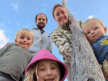 A happy family of five poses outdoors, smiling down at the camera near a tall wooden post, with a bright blue sky in the background. Playful and joyful.
