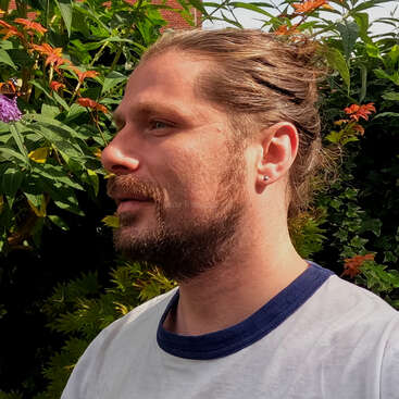 Um homem com barba e cabelo amarrado para trás está do lado de fora, sob a luz do sol. Ele veste uma camiseta branca com detalhes azuis, cercado por flores e folhagens verdes exuberantes.