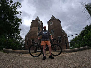 Um homem está parado com sua bicicleta em frente a um edifício histórico de tijolos com duas torres, sob um céu nublado, cercado por árvores e caminhos de pedra.