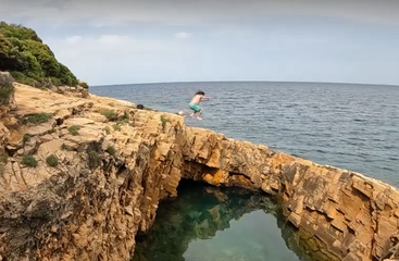 Uma pessoa pula de um penhasco rochoso acidentado em direção ao mar, com água azul clara abaixo e vegetação visível ao fundo sob um céu calmo.