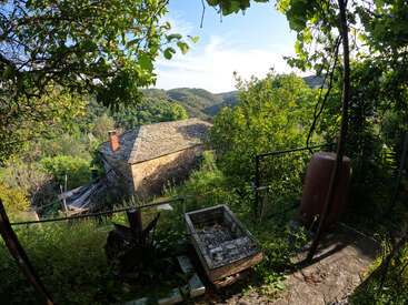 Uma casa rústica de pedra com telhado de ardósia aninhada entre árvores e colinas verdejantes, vista de um jardim com uma pia de concreto e um barril de água.