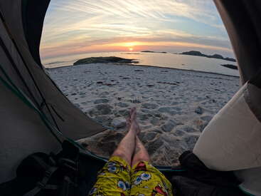 Uma pessoa relaxa em uma barraca em uma praia de areia, usando shorts do Bob Esponja, assistindo a um sereno pôr do sol sobre o oceano com ilhas rochosas distantes. Atmosfera tranquila.
