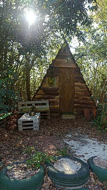 The image depicts a small wooden cabin nestled among trees, featuring a bench, table, and tires repurposed as planters, with sunlight filtering through the foliage.