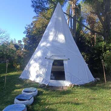 The image depicts a white teepee with an open door, situated in a grassy area surrounded by trees, with a clear blue sky in the background.
