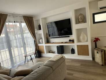 Modern living room with built-in shelves, flat-screen TV, decorative objects, light wood flooring, beige curtains, cozy sofa, side table, and natural sunlight through large windows.