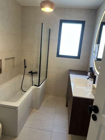 This modern bathroom features a white bathtub with black fixtures, a glass partition, large tiles, vanity with a sink, bright window, and a round mirror.