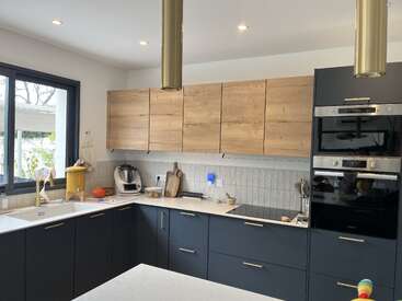 This modern kitchen features sleek dark cabinets, wooden upper cupboards, built-in appliances, a large window, marble countertops, pendant lighting, and various kitchen gadgets for convenience.