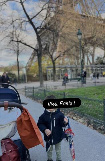 A young child stands near a stroller with an umbrella in hand, under the Eiffel Tower in Paris. The image encourages viewers to "Visit Paris!"