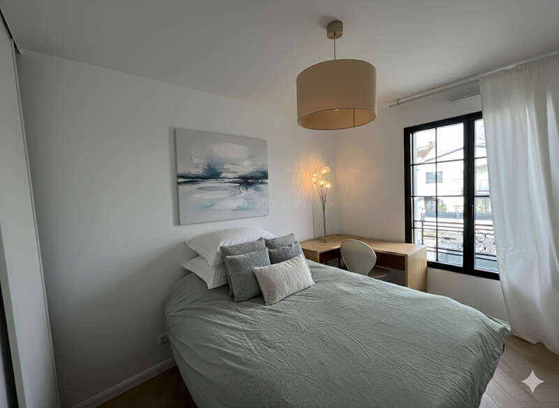 This cozy bedroom features a neatly made bed with multiple pillows, modern art, a stylish desk by the window, soft lighting, and minimalist, contemporary decor.