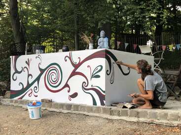 Uma mulher está pintando um mural em uma parede branca do lado de fora, usando um pincel e um balde de tinta por perto. O mural apresenta padrões em redemoinho em verde e vermelho.